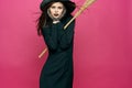 woman in black suit witch broomstick posing pink background