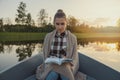 woman with blanket on shoulders sitting in a row boat on the lake and read a book at sunset Royalty Free Stock Photo