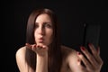 Woman blowing lips sending air kisses over palm, taking selfie to mobile phone on black background