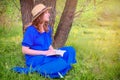 A woman in a blue dress reading a book sitting near a tree. Girl in a straw hat in the summer forest Royalty Free Stock Photo