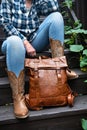woman in blue jeans and cowboy boots sits on the steps and holds a leather backpack. Close-up Royalty Free Stock Photo