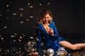 A woman in a blue sequined dress is smiling sitting on the floor with confetti on a black background