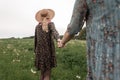 Woman in blue stylish dress holds girlfriends hand in straw vintage hat in black dress along field with green grass. Two sister in Royalty Free Stock Photo