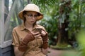 Woman botanist dressed in safari style in greenhouse. Naturalist in khaki clothes, leaver gloves resting and reading a book
