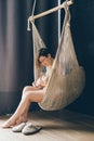Woman is breastfeeding a newborn baby sitting in a cozy wicker hammock suspended in the interior of the apartment on a dark backgr