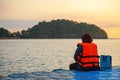 A woman in a bright orange life jacket sits on a pontoon pier early in the morning during sunrise. Dangling legs in the water and Royalty Free Stock Photo