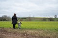 Woman with brown curly hair standing next to her akita inu dog with gray fur in front of an agricultural field, rear view, Royalty Free Stock Photo