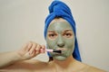 A woman with a brush applies a clay mask to her face with blue towel on her head