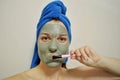 A woman with a brush applies a clay mask to her face with blue towel on her head