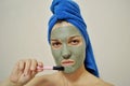 A woman with a brush applies a clay mask to her face with blue towel on her head