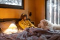 Woman cares her dog while lying in bed in tiny bedroom of wooden cabin on nature