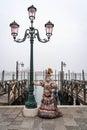 Lady in masquerade costume a mirror in her hand, stands leaning on a lamppost at Venice carnival, San Marco square, Italy