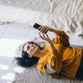 Woman in casual clothing lying down the floor in her livingroom and browsing internet on her smartphone. Leisure time of single