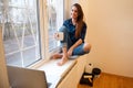 Woman in casual denim clothes sitting on the window sill and drinking coffee Royalty Free Stock Photo