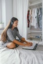 Woman in casual gray dress with headphones using laptop sitting on the bed in the bedroom at home, freelance and home study Royalty Free Stock Photo