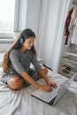 Woman in casual gray dress with headphones using laptop sitting on the bed in the bedroom at home Royalty Free Stock Photo