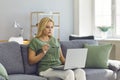 Focused freelancer woman working on laptop sitting on sofa in living room of her home.