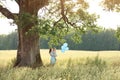 A woman with a child with blue balloons under a huge old oak tree with mighty branches.