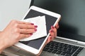 Woman cleaning tablet device with cotton paper tissue, wiping black screen, closeup detail pink color fingernails. Blurred laptop