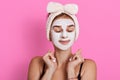 Woman with closed eyes and white facial mask on face, wearing sleeveless t shirt and hair band, standing against pink background, Royalty Free Stock Photo