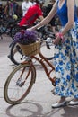 A woman in a colored skirt holds the wheel of a bicycle. Basket of flowers. Frame part