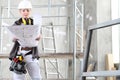 Woman construction worker builder looking at bluprint, wearing helmet, hearing protection headphones and tools belt bag in Royalty Free Stock Photo