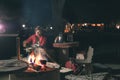 Woman cooking with fire wood and braai equipment by night. Tent and chairs in the foreground. Adventures in african national parks