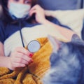 A woman with a coronavirus in a medical mask plays with a cat while lying in bed. Girl with a stethoscope and pet on the couch