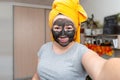 A woman with a cosmetic mask on her face made of clay, a towel on her head makes a selfie