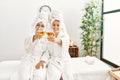 Woman couple wearing bathrobe toasting with champagne sitting on massage board at beauty center