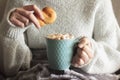 Woman covered with blanket holding biscuit and mug of hot drink with whipped cream