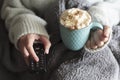 Woman covered with blanket holding tv remote and mug of hot drink with whipped cream