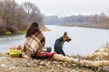 A woman covered with warm plaid is sitting backside at tha bank river with a dog by her side