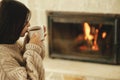 Woman in cozy sweater drinking cup of warm tea at fireplace in rustic room. Heating house in winter with wood burning stove. Young Royalty Free Stock Photo