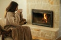 Woman in cozy sweater drinking cup of warm tea at fireplace in rustic room. Heating house in winter with wood burning stove. Young Royalty Free Stock Photo