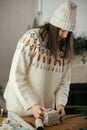 Woman in cozy sweater and hat wrapping christmas gift in paper on wooden table with festive decorations in decorated scandinavian Royalty Free Stock Photo