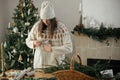 Woman in cozy sweater and hat wrapping christmas gift in paper on wooden table with festive decorations in decorated scandinavian Royalty Free Stock Photo