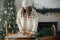 Woman in cozy sweater and hat wrapping christmas gift in paper on wooden table with festive decorations in decorated scandinavian Royalty Free Stock Photo