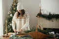 Woman in cozy sweater and hat wrapping christmas gift in paper on wooden table with festive decorations in decorated scandinavian Royalty Free Stock Photo