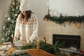 Woman in cozy sweater and hat wrapping christmas gift in paper on wooden table with festive decorations in decorated scandinavian Royalty Free Stock Photo