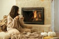 Woman in cozy sweater holding cup of warm tea at fireplace, autumn hygge. Heating house with wood burning stove. Young stylish