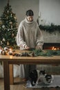 Woman in cozy sweater making Christmas rustic wreath on wooden table with cute cat in festive living room with fireplace. Winter Royalty Free Stock Photo