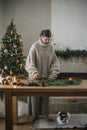 Woman in cozy sweater making Christmas rustic wreath on wooden table with cute cat in festive living room with fireplace. Winter Royalty Free Stock Photo