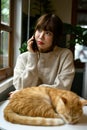 A woman in cozy sweater talking on the phone while her cat is sleeping on a table