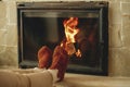 Woman in cozy wool socks warming up feet at fireplace in rustic room. Heating house in winter with wood burning stove. Young