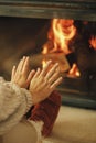 Woman in cozy wool socks warming up feet and hands at fireplace in rustic room. Heating house in winter with wood burning stove.