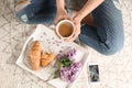 Woman with cup of coffee and tray with tasty croissants and beautiful blossoming lilac on carpet