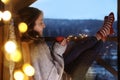 Woman with cup of hot beverage and Christmas lights resting on balcony