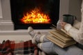 Woman with cup of hot drink and book resting near fireplace at home, closeup Royalty Free Stock Photo