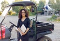 Woman with curly hair in black dress holding in hands a laptope, poses near vehicle and smiling. Small bussines content Royalty Free Stock Photo